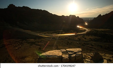 Traffic At Night Driving On Windy Road Through The Desert In Utah As The Landscape Is Lit Up By The Full Moon.
