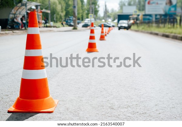 Traffic safety\
cones on asphalt of road. Selective focus on striped orange-white\
warning signs, cars\
bokeh.
