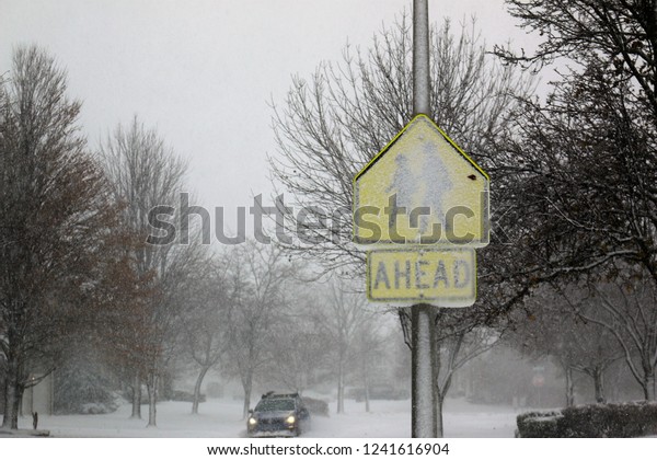 Traffic sign of a school, covered in sleet,\
during a snow blizzard, while the surrounding is covered in several\
inches of snow and a car is approaching in the background. Concept\
of closed schools.