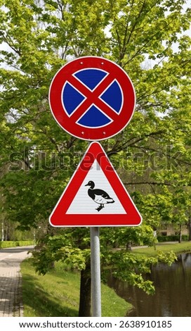 Traffic signs by a riverside path: a no parking symbol above a duck crossing warning, set against a lush green background, alerting drivers to wildlife presence near water.