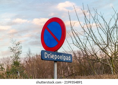 Traffic signs: Parking prohibited, street name Driepoelpad against cloudy sky, round plate with red and blue colors, bare tree branches background, Sittard-Geleen, South Limburg, the Netherlands - Powered by Shutterstock