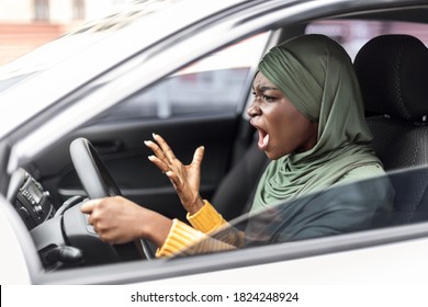 Traffic Stress. Angry black muslim female in hijab driving car and shouting at somebody, emotionally reacting to accident or road jam, holding steering wheel with displeased face expression, side view