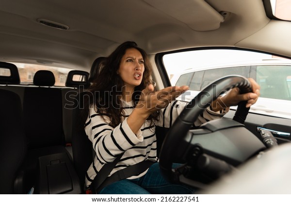 Traffic Stress. Angry female driving car shouting at\
somebody, emotionally reacting to accident or road jam holding\
steering wheel with displeased face expression, waving hand,\
windshield view
