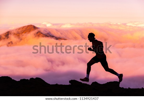 Trail\
runner athlete man running in nature landscape. Silhouette of male\
person training on mountains in cold weather with pink clouds at\
sunset. Amazing mountain peaks in the\
background.