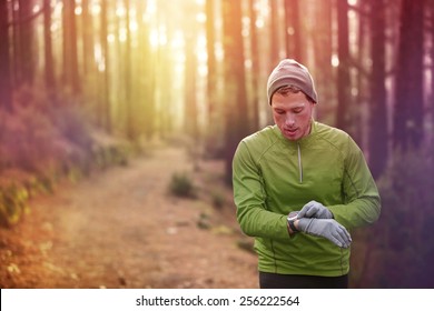 Trail running runner looking at heart rate monitor watch running in forest wearing warm jacket sportswear, hat and gloves. Male jogger running training in woods.