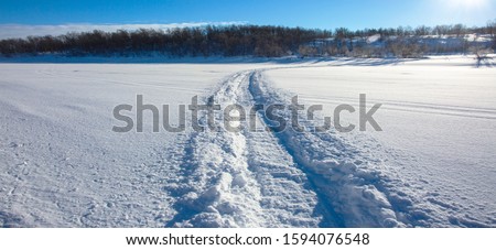 Trail from a snowmobile on a frozen and snowy river bed. Panoramic view of the river bank. Norway