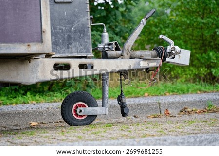 A trailer front shows a small support wheel jack and metal hitch head ready to couple to a car, the steel parts and chain links sit on rough ground like a parked unit waiting for motion.