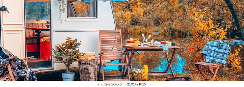 Trailer Of Mobile Home Or Recreational Vehicle Stands On Shore Of Pond In Camping In Autumn Near Table Set, Concept Of Family Local Travel In Native Country On Caravan Or Camper Van And Camping Life