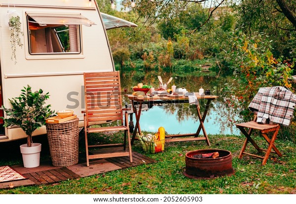 trailer of mobile home or recreational vehicle\
stands on shore of pond in camping in autumn near table set,\
concept of family local travel in native country on caravan or\
camper van and camping\
life