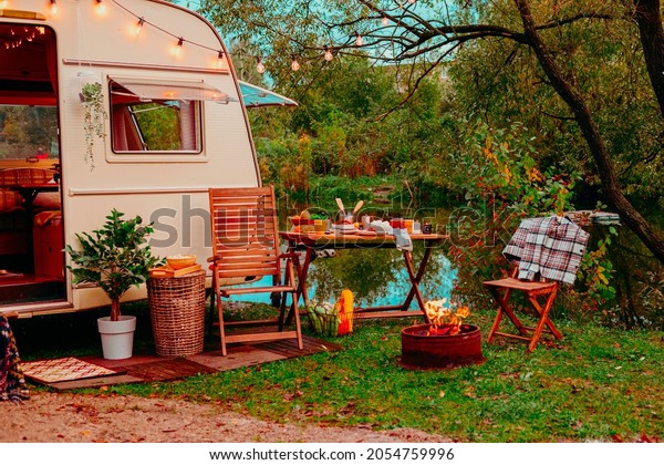 trailer of mobile home or recreational vehicle\
stands on shore of pond in camping in autumn near table set,\
concept of family local travel in native country on caravan or\
camper van and camping\
life