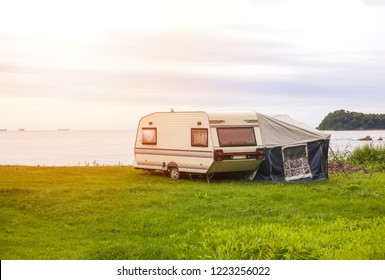 Trailer Motor Home And A Tent On The Grassy Part Of The Beach At Sunset. Leisure Mobile Camping Home For Tourists Overlooking The Blue Sea And Cape. Adventure Relaxing Travel On Caravan Van