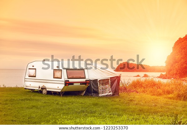Trailer motor home and a tent on the grassy part of\
the beach at sunset. Leisure mobile camping home for tourists\
overlooking the blue sea and cape. Adventure relaxing travel on\
caravan van