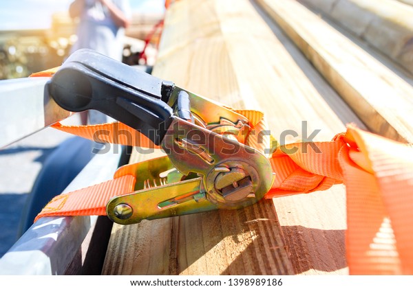 Trailer strop or strap in orange\
nylon and metal tie down with wood and trailer. Object helping for\
holding stuff , storage and transport for safty and\
security.