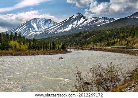 Train crossing the Alaskan frontier back country, passing mountain peaks and flowing rivers.