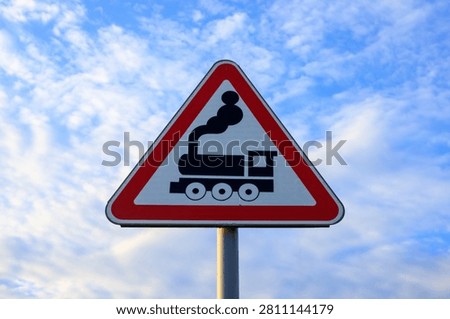 Train crossing warning sign. Triangular metal plate with locomotive symbol and red border under blue sky with light clouds outdoor