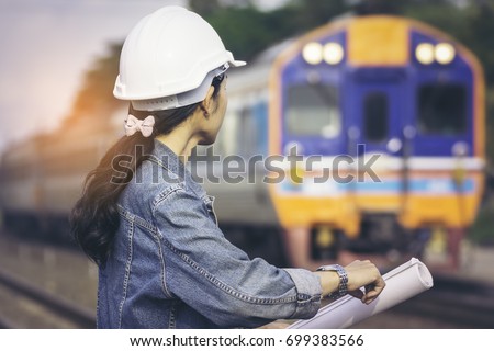 Train engineers beautiful girl wearing jeans and a white hard hat looking at the clock and checking train times to and from the station. A train is passing.