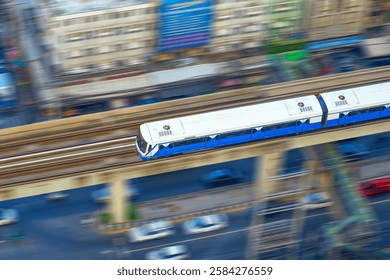 A train glides along elevated tracks above a busy city street. Colorful buildings and vehicles can be seen below, showcasing urban life during a sunny day.