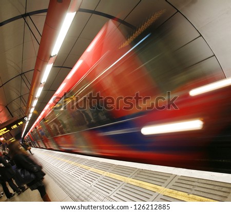 Train, motion blurred, passing through a DLR, Docklands Light Railway, station and people are present at platform,