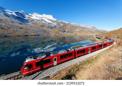 A train of Rhaetian Railway travels along Lago Bianco in Bernina Pass on a brisk autumn day with mountain peaks covered by snow and reflected in the water under blue clear sky, in Grisons, Switzerland - Shutterstock ID 2139513271