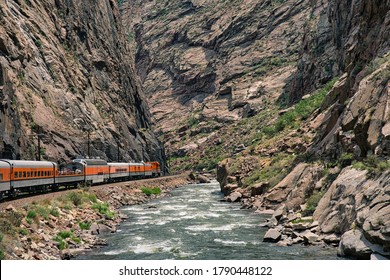 Train Riding Deep In The Royal Gorge Beside The Arkansas River In Colorado. Orange Engine Beside Rushing Blue Water With Rocky Cliffs All Around