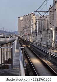 Train station platform at dusk with empty railway tracks, modern station architecture, and illuminated residential towers in the background under twilight sky.
