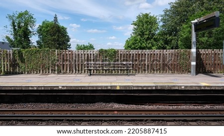 Train station platform with a wooden bench and fence. Trees and blue sky with white clouds in the background.