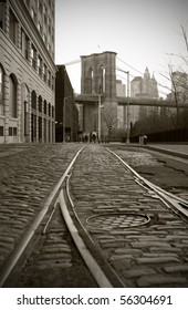 Train Tracks On Cobblestone Road In Dumbo Area Of Brooklyn, Brooklyn Bridge Tower In Background, Black And White.