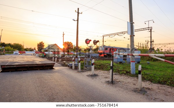 Train tracks, Red traffic lights and stop signs at a\
protected rural railway crossing barrier in the rays of the setting\
sun.