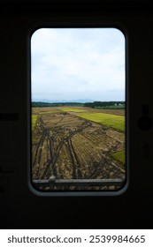 Train window view of patchy fields with green and brown hues, distant trees, and a cloudy sky.