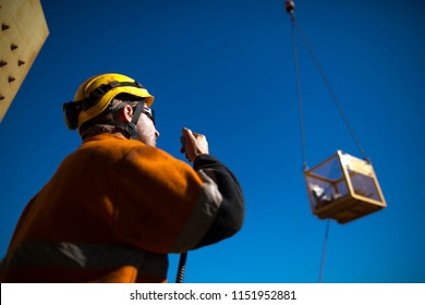 Trained Rigger Wearing Safety Helmet, Orange Long Sleeve Shirt Using Two Way Radio Communicating With Crane Operator While Load Is Being Lifted In The Afternoon Construction Site Perth, Australia 