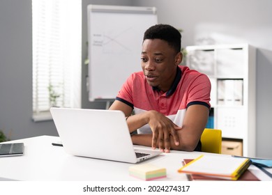 Trainee in office sits at desk in front of laptop monitor in background whiteboard, young dark-skinned man talks online with clients, co-workers, uses webcam, video call