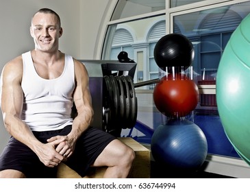 Trainer With Healthy And Strong Body, Inside Room Of A Luxury Gym, Heavy Exercise Tools Near Wall, A Body Builder Sitting On A Bench, He Is Smilling And Relaxing.