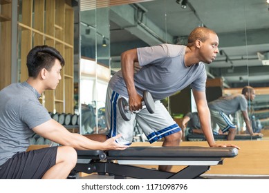 Trainer Helping Man Lifting Dumbbell In Gym. Young Black Guy Training With Personal Trainer. Bodybuilding Concept. Side View.