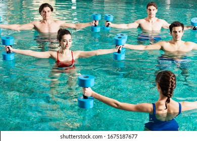 Trainer holding barbells while exercising with group of young people in swimming pool