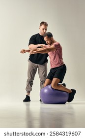 Trainer instructs man balancing on stability ball, engaging core muscles and coordination for post-training rehabilitation against white studio background. Concept of sport, recovery, healthcare
