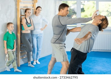 Trainer and man in group self-defense classes practicing sparring technique of blowing to chin in gym, in background family watches of performing technique