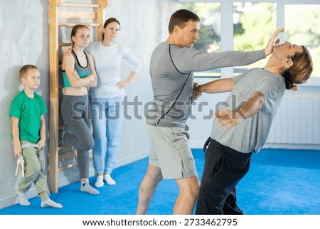 Trainer and man in group self-defense classes practicing sparring technique of blowing to chin in gym, in background family watches of performing technique