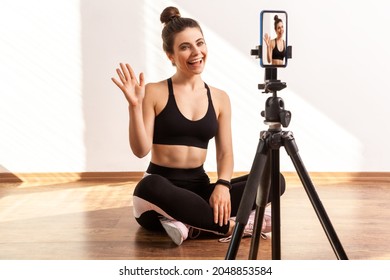 Trainer Saying Hello Or Bye, Recording Online Tutorial Of Fitness Exercises, Using Hone On Tripod, Wearing Black Sports Top And Tights. Full Length Studio Shot Illuminated By Sunlight From Window.