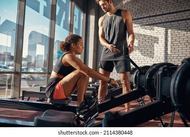 Trainer showing black girl sport indicators on training apparatus screen while she exercising in gym. Healthy lifestyle. Smiling woman and caucasian man wearing sportswear. Fitness center in sunny day - Shutterstock ID 2114380448