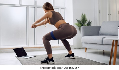 A training adult is looking at the computer while doing squats. There is a gray sofa near her. She has black sneakers. She is also wearing brown clothes. Her hands is locked. Her computer is white.