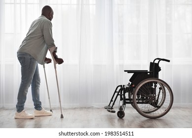 Training After Injury, Recovery At Home, Exercise In Rehabilitation Clinic. Tense Adult African American Guy Disabled Walks With Crutches To Wheelchair On Large Window Background In Room Interior