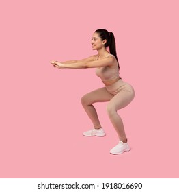 Training Buns, Hips And Thighs. Positive Fit Young Lady In Sportswear Doing Deep Squat Exercise With Straight Outstreched Arms During Work Out Practice, Isolated Over Pink Studio Background