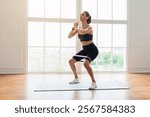 Training Concept. Confident determined sportswoman squatting with elastic resistance band in fitness studio or at home in living room doing leg and buttocks workout, listening to music in earbuds