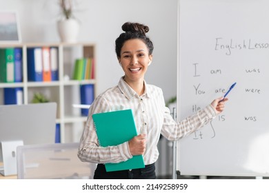 Training concept. Happy female tutor teaching English language, pointing at grammar rules on board and smiling at camera, holding clipboard in office interior - Shutterstock ID 2149258959