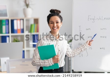Training concept. Happy female tutor teaching English language, pointing at grammar rules on board and smiling at camera, holding clipboard in office interior