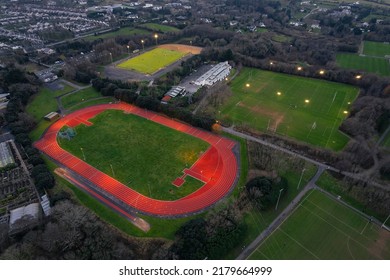 Training field with tall goal posts for Irish National sport, camogie, hurling, rugby, Gaelic football. Running track with red surface. Illuminated at dusk time. Aerial view.