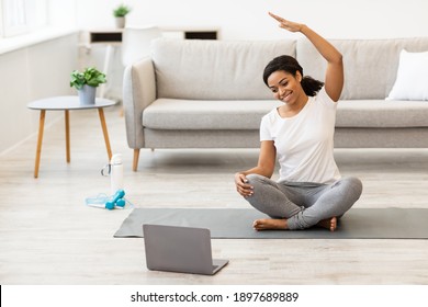 Training At Home. Active young black woman practicing yoga in side bend pose, stretching arm and watching tutorial training on laptop in the morning, sitting on mat in bedroom, free space - Powered by Shutterstock