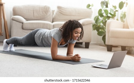 Training At Home. Fit Black Woman Doing Yoga Plank While Watching Online Tutorial On Laptop, Exercising In Living Room, Free Space, Panorama