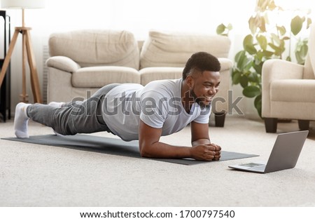 Training At Home. Sporty man doing yoga plank while watching online tutorial on laptop, exercising in living room, free space