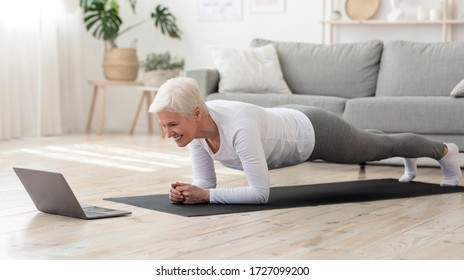 Training At Home. Sporty senior woman doing yoga plank while watching online tutorial on laptop, exercising in living room, free space, panorama